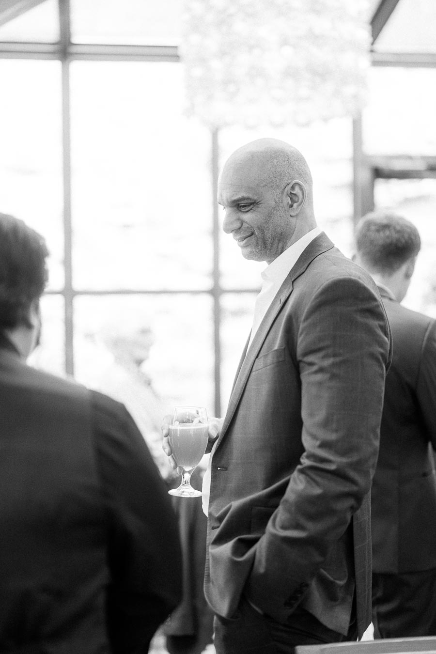 Man in a suit smiles while holding a drink during an indoor wedding reception gathering.