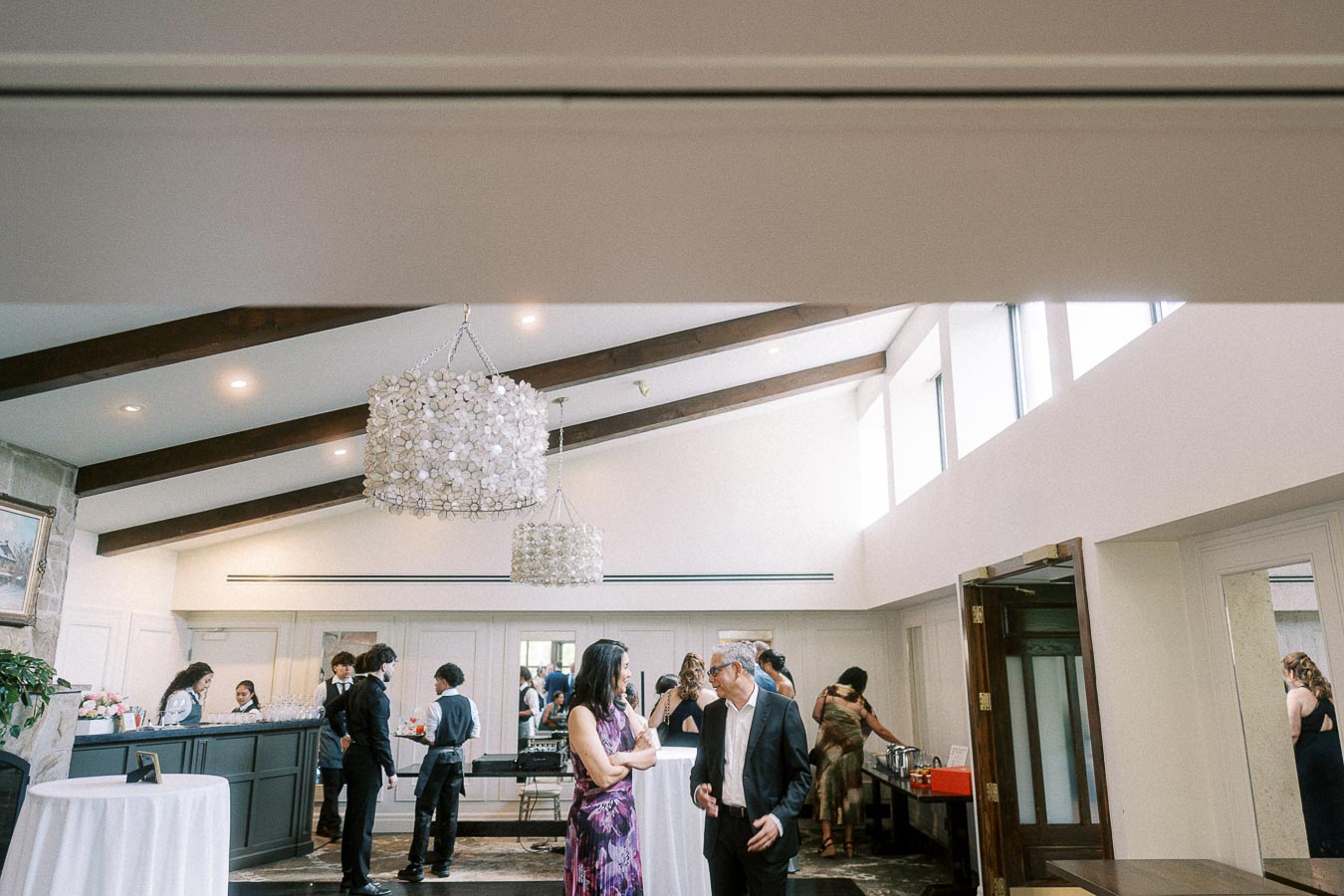 Guests mingle in a bright Ancaster Mill reception room with chandeliers, beams, bar, and buffet setup.