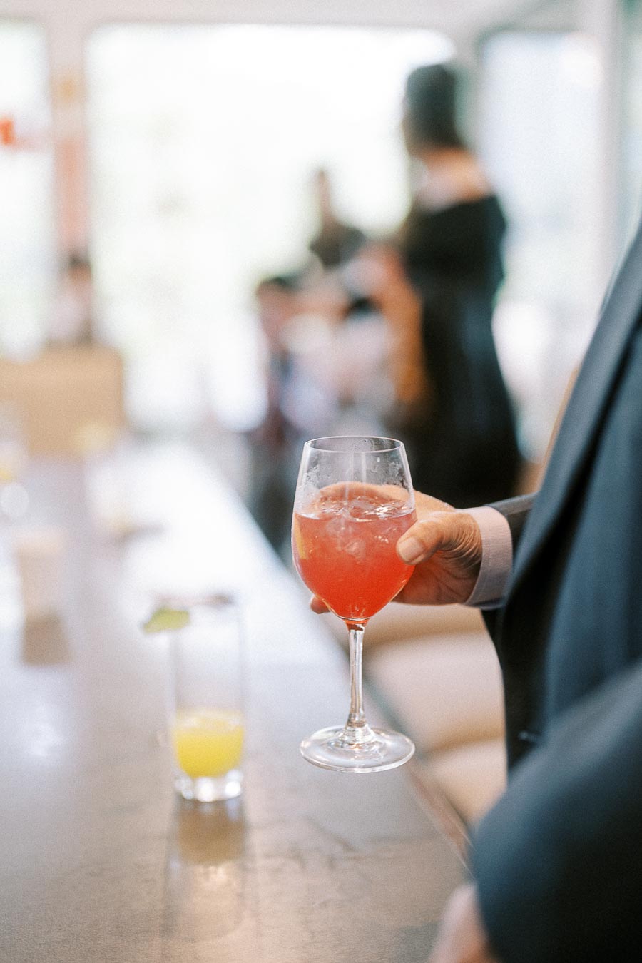Hand holding a stemmed glass of pink cocktail indoors, with blurred wedding guests in the background.