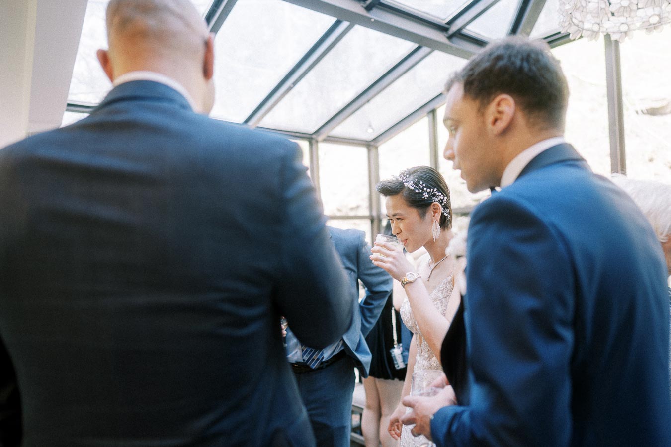 Bride in a beaded gown drinks from a glass among suited guests in a bright glass-roofed room.
