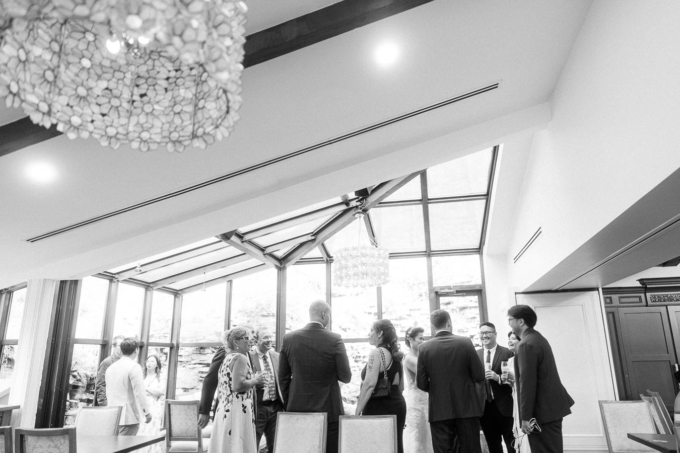 Wedding guests mingle in a bright glass-walled room with chandeliers at Ancaster Mill.