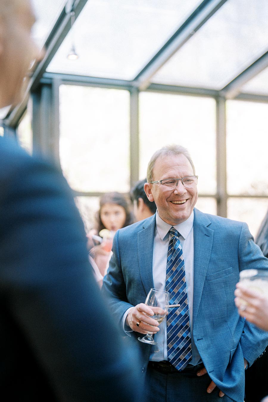 Smiling man in a blue suit holds a drink while chatting with guests in a bright glass-walled wedding space.