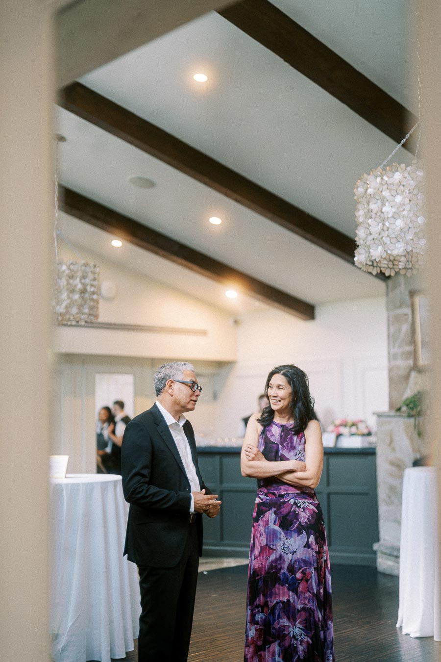 Two wedding guests talk inside Ancaster Mill reception room beneath wood beams and shell chandeliers.