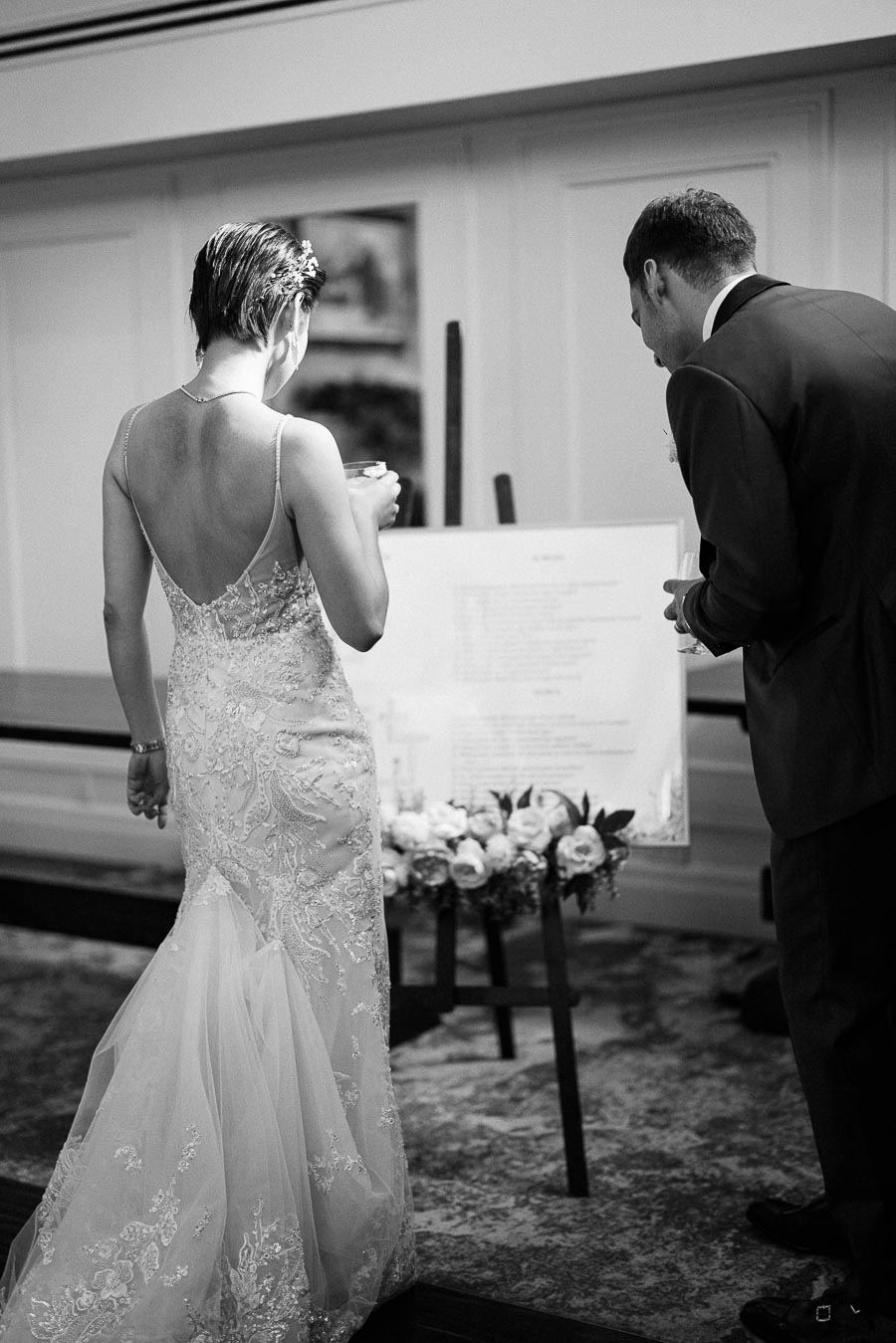 Bride in a lace gown and groom in a dark suit look at a seating chart during their wedding reception.