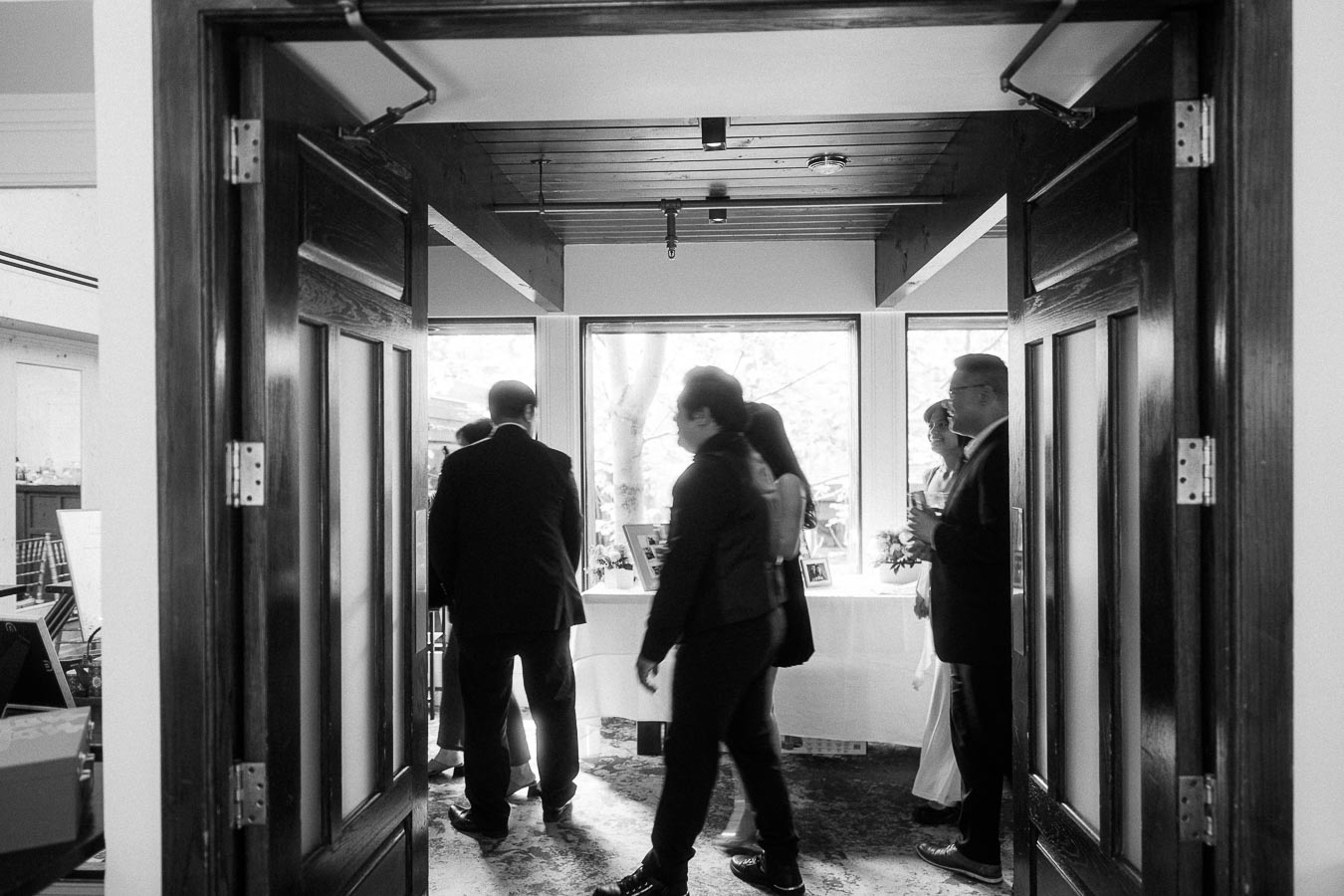 Guests in formalwear mingle near open wooden doors and a bright window during an indoor wedding reception.