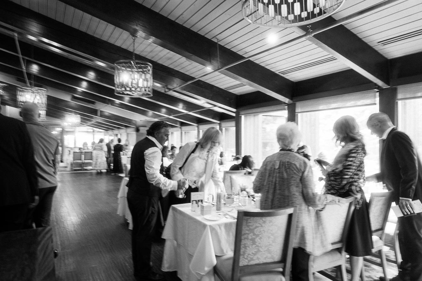 Black-and-white candid of wedding guests greeting each other beside reception tables in a bright dining room.