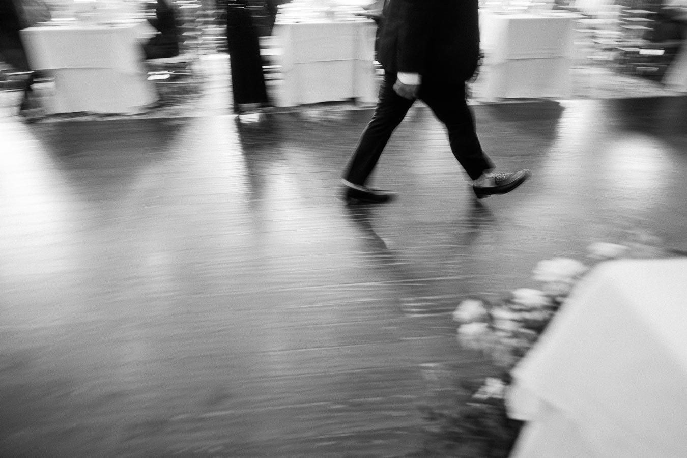 Blurred black-and-white view of a suited guest walking across a reception dance floor.