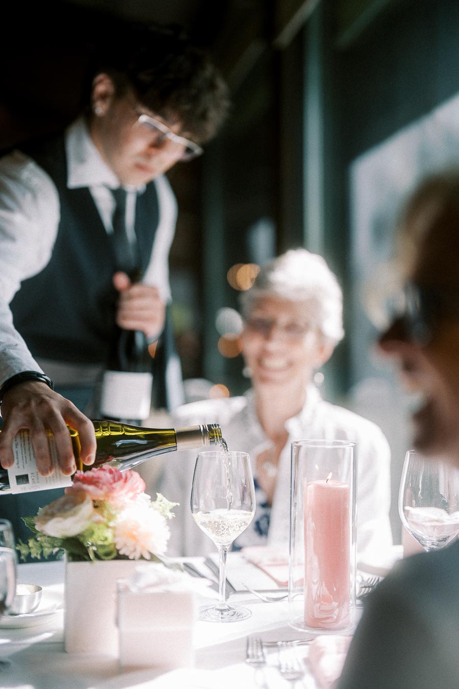Server pours white wine into a guest's glass at a sunlit wedding reception table with pink candles and flowers.