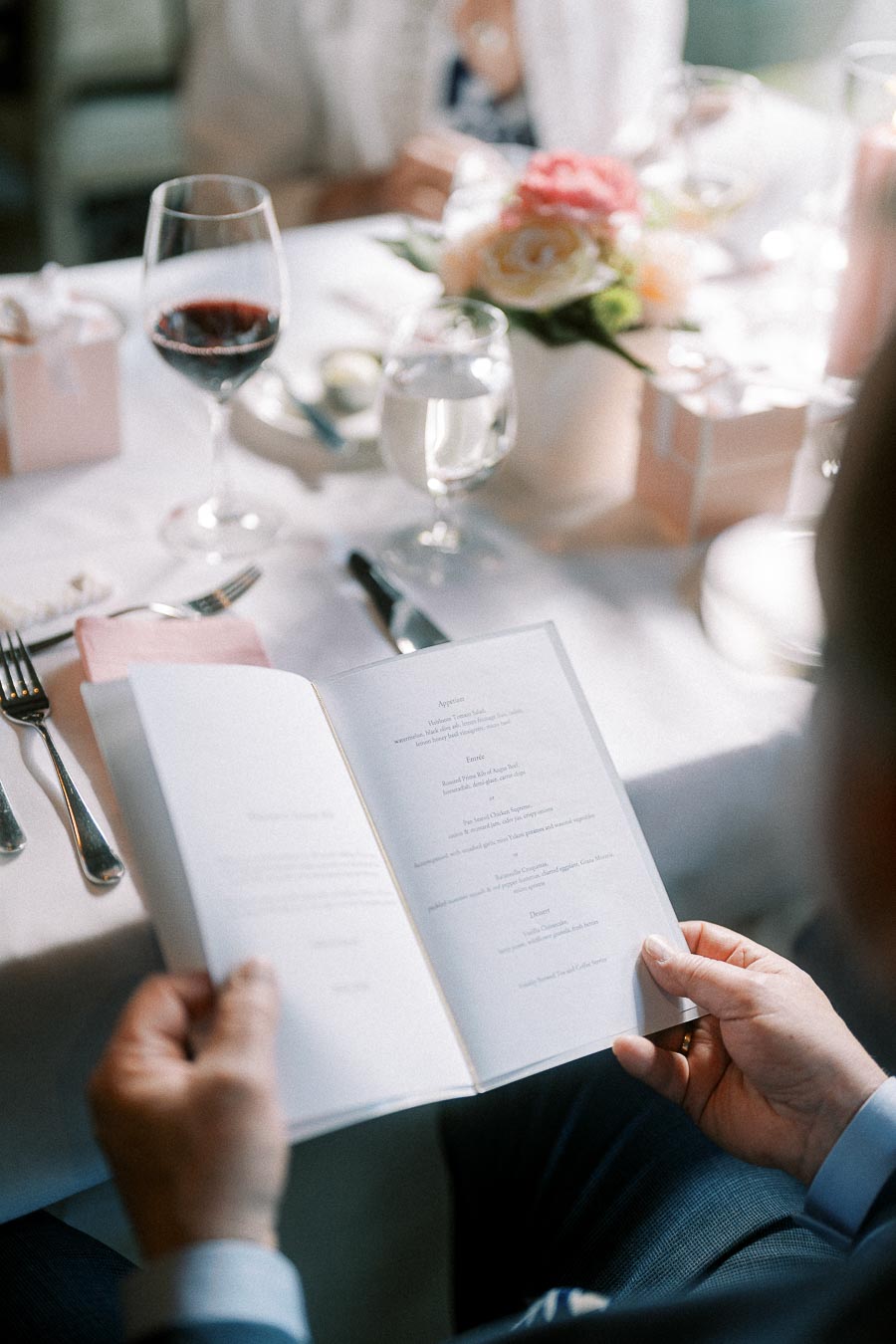 Guest reads a wedding reception menu at a table with wine, water, blush napkins, and a floral centerpiece.