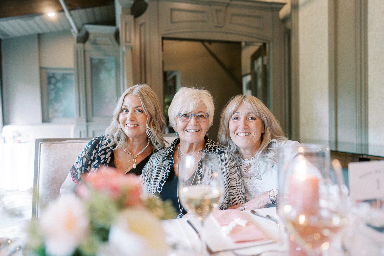 Three smiling women seated together at a wedding reception table with flowers, candles and wine glasses