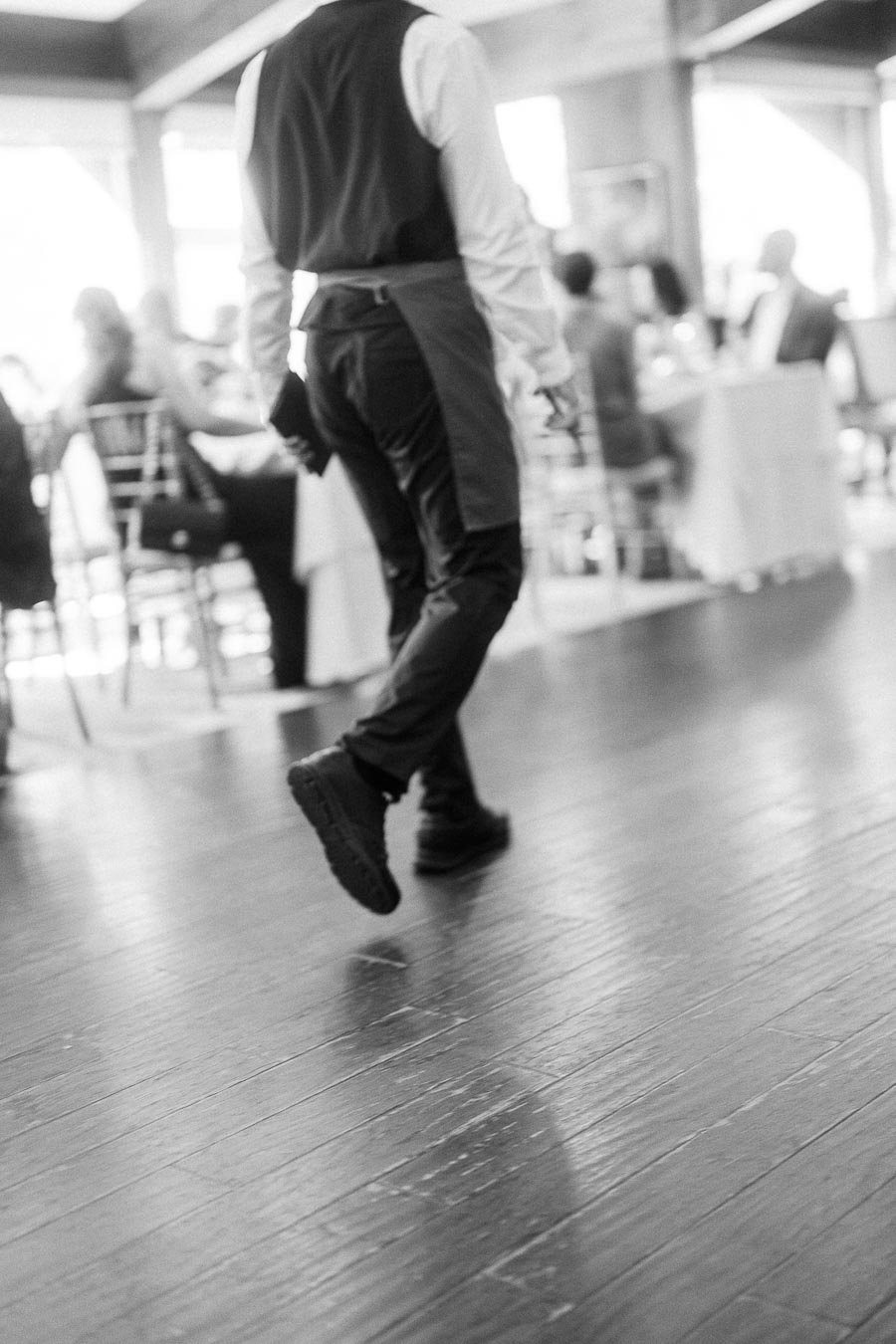 Black-and-white view of a server walking across a polished reception floor with seated guests in the background.