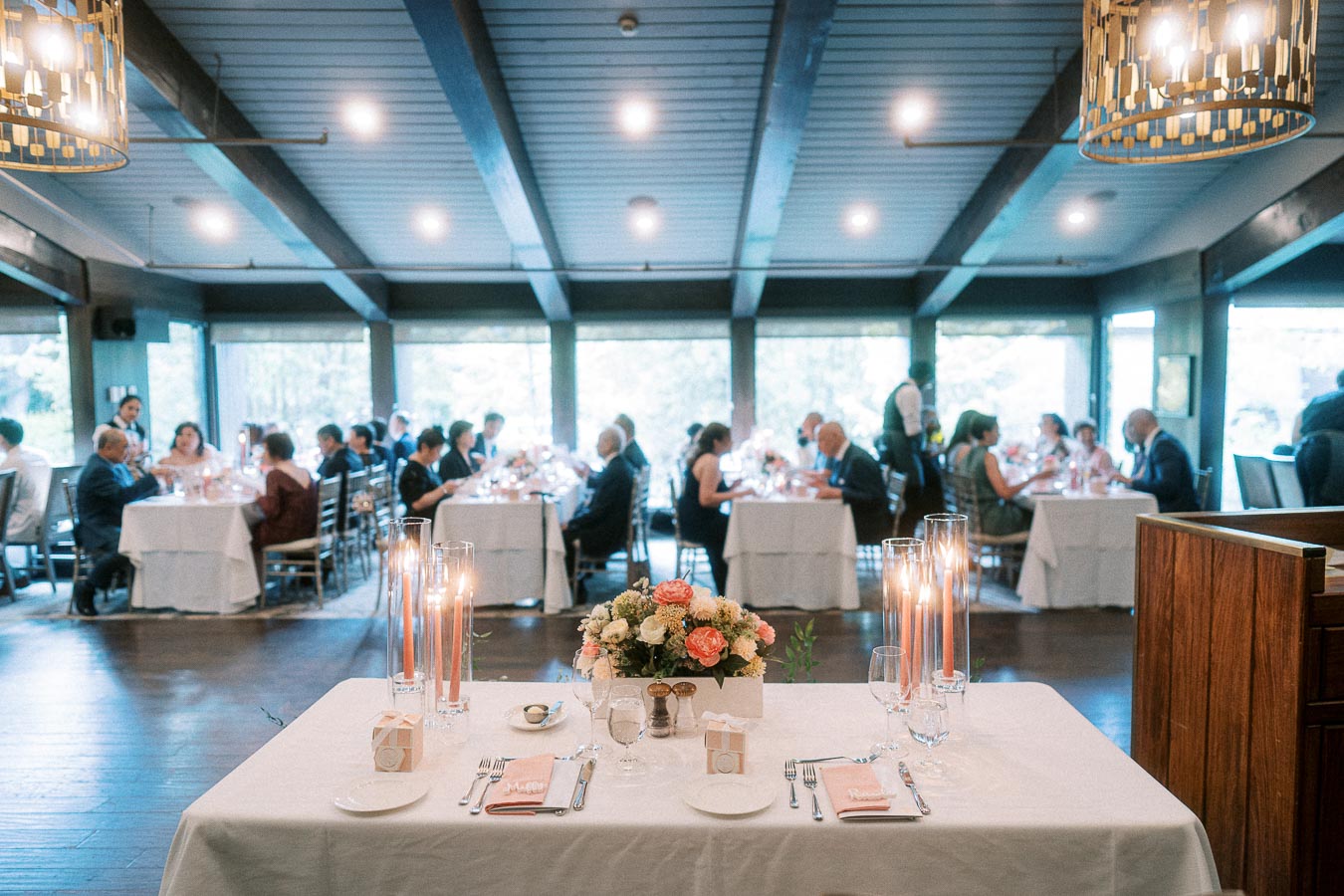 Head table with candles and flowers set before seated wedding guests in a bright reception room.