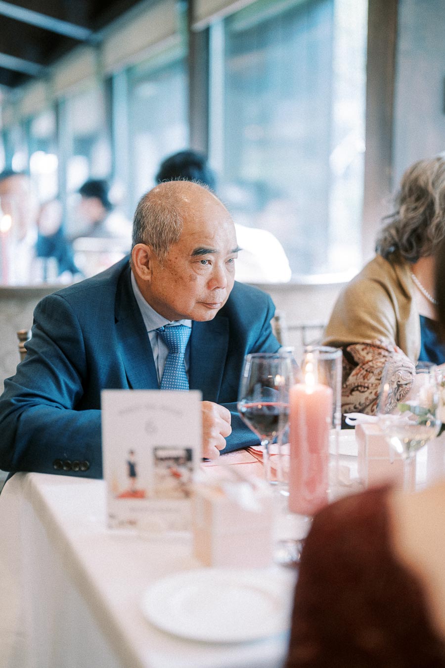 Man in a blue suit seated at a wedding reception table with wine glasses and a pink candle.