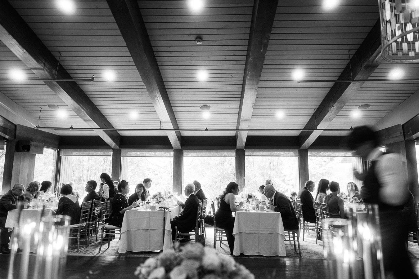 Guests seated at round tables in a windowed reception room with timber beams and candles in the foreground.
