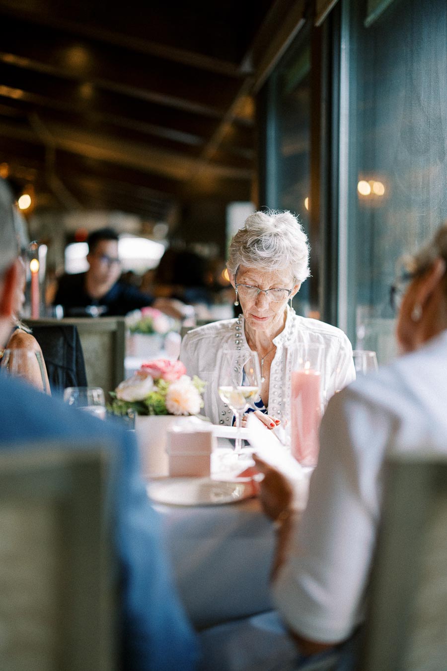 Older woman seated at a reception table reading a card beside pink flowers and candlelight.