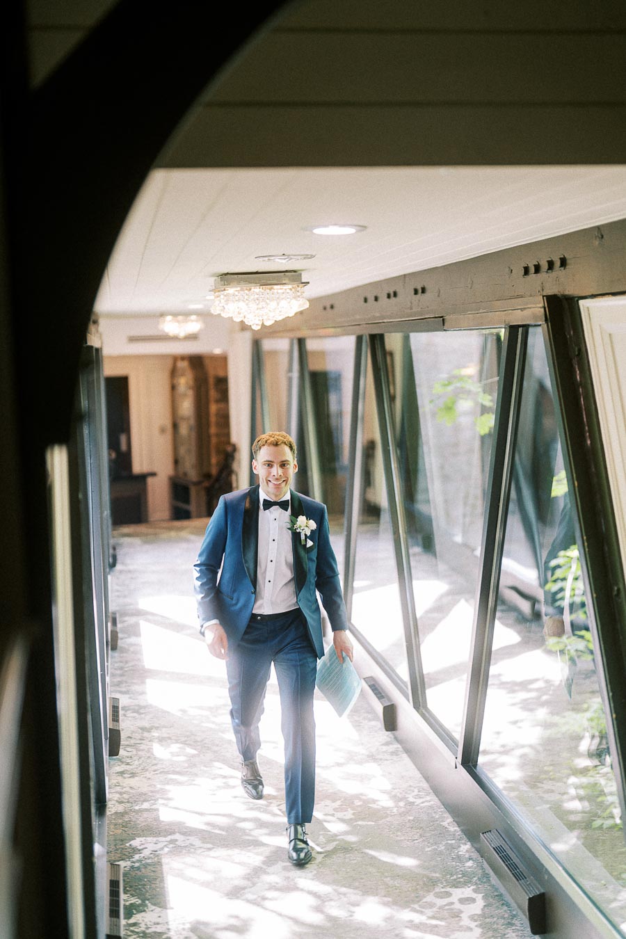 Groom in a blue tuxedo walks through a bright glass hallway at Ancaster Mill holding papers.