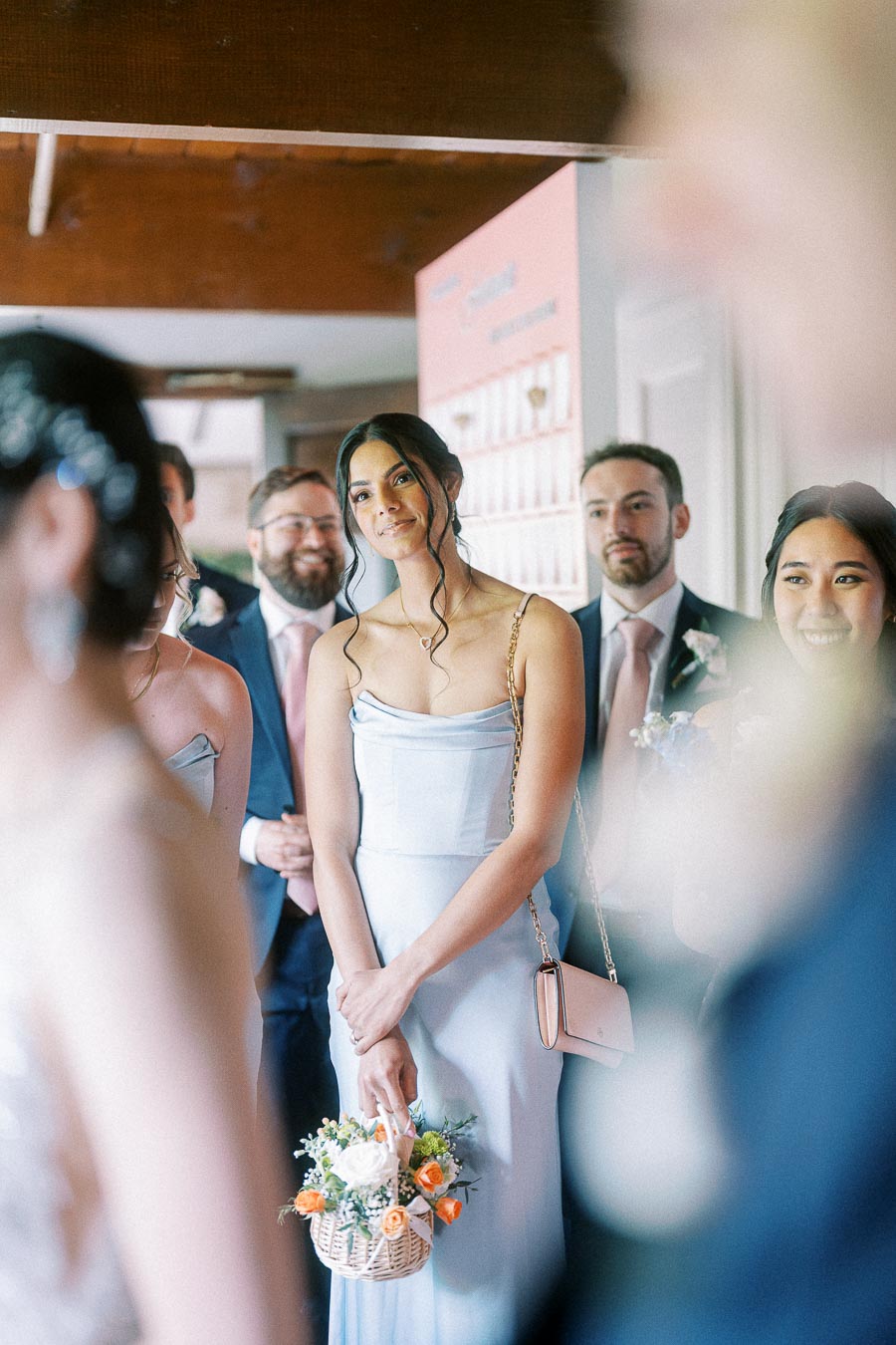 Wedding guest in a pale blue dress holds a small flower basket among smiling guests indoors.