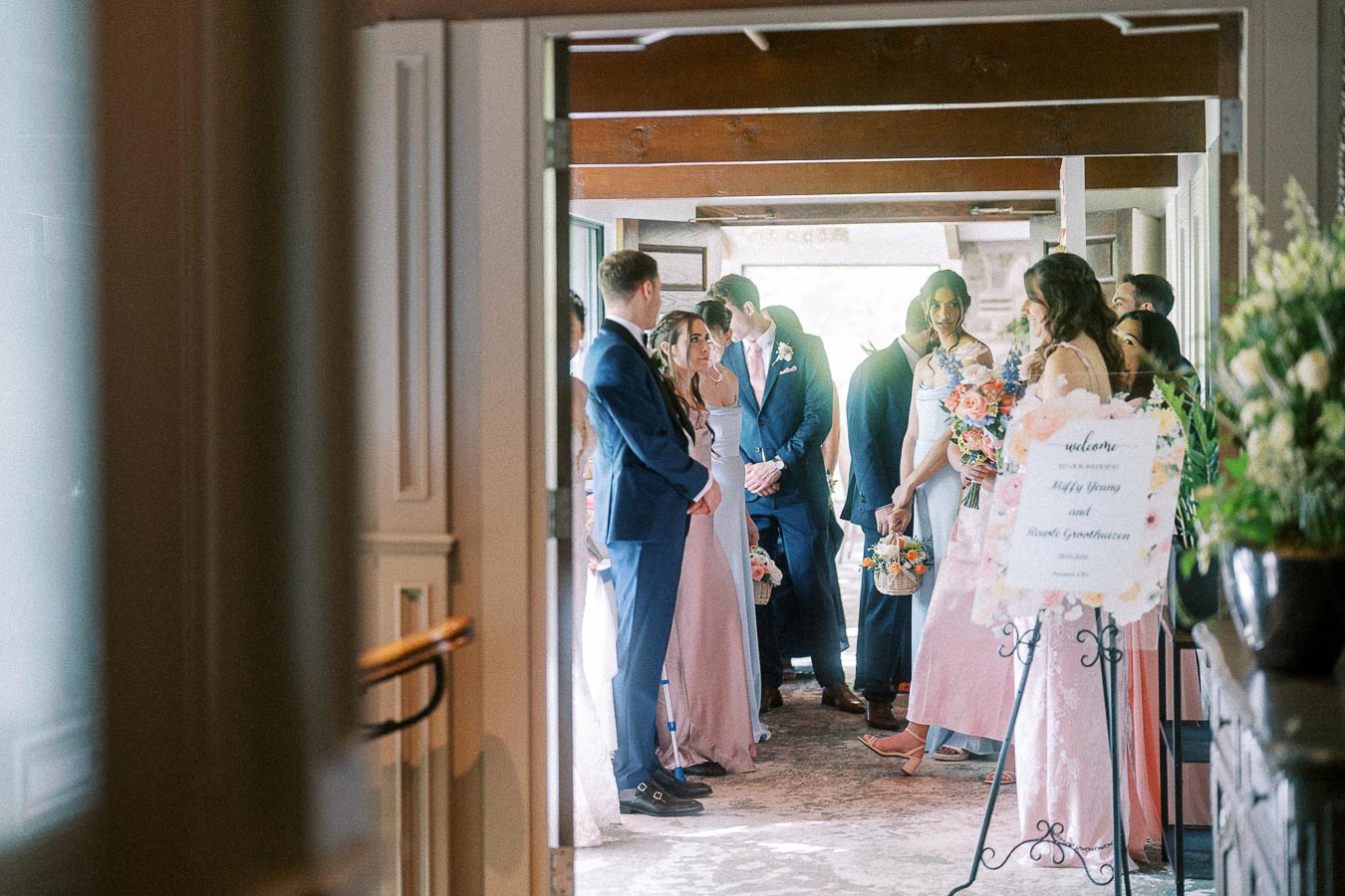 Wedding party in pastel pink dresses and navy suits gathers by a welcome sign inside Ancaster Mill.