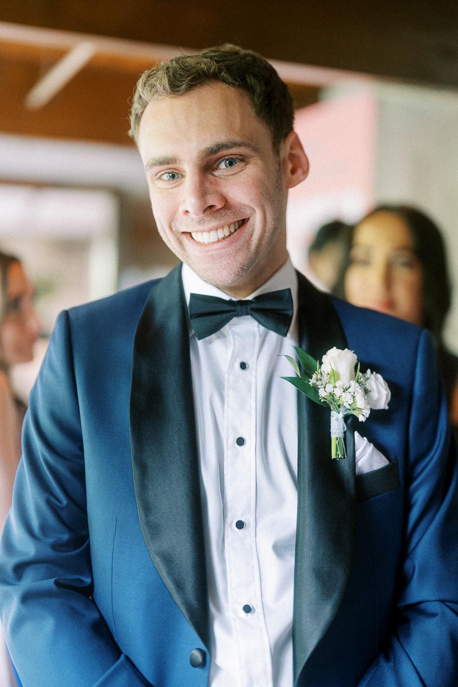 Smiling groom in a blue tuxedo with black bow tie and white boutonniere, standing indoors with guests behind him.