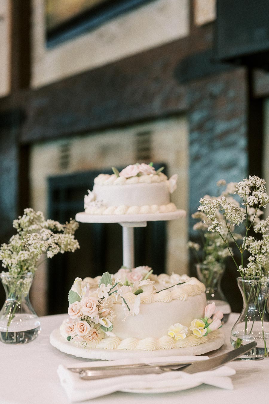 Two-tier white wedding cake with pale flowers and baby’s breath vases on a reception table.