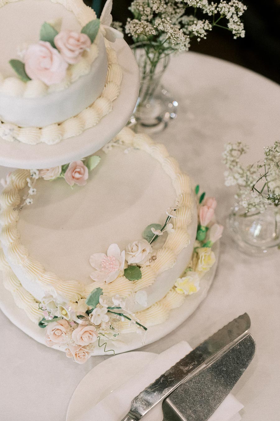 Two-tier white wedding cake with pastel sugar flowers, piped icing, baby’s breath, and serving knife on a table