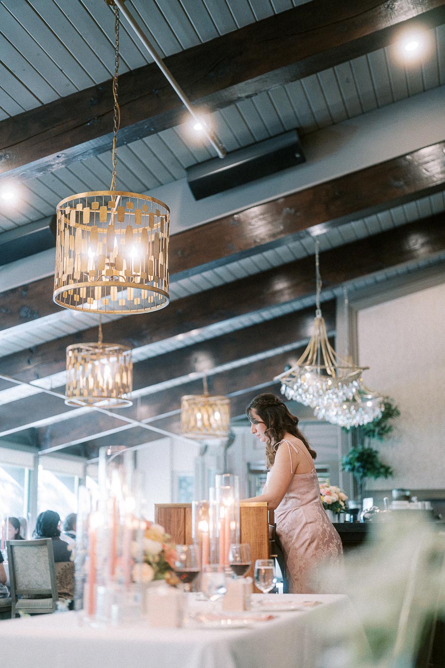 Woman in a satin dress speaks at a wooden podium beneath chandeliers in an elegant wedding reception room.