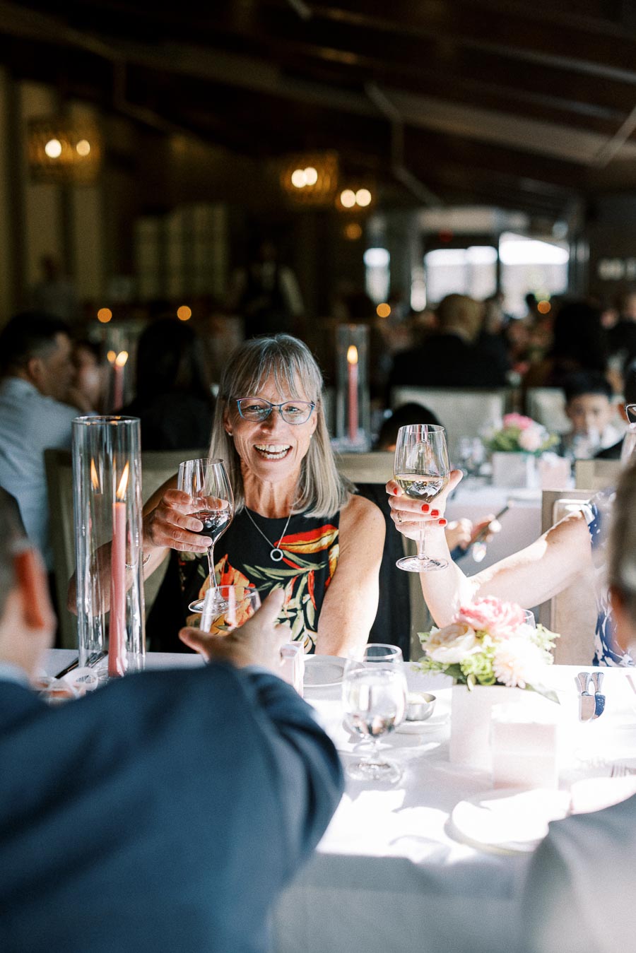 Smiling wedding guest raises a wine glass at a candlelit reception table with other guests around her.