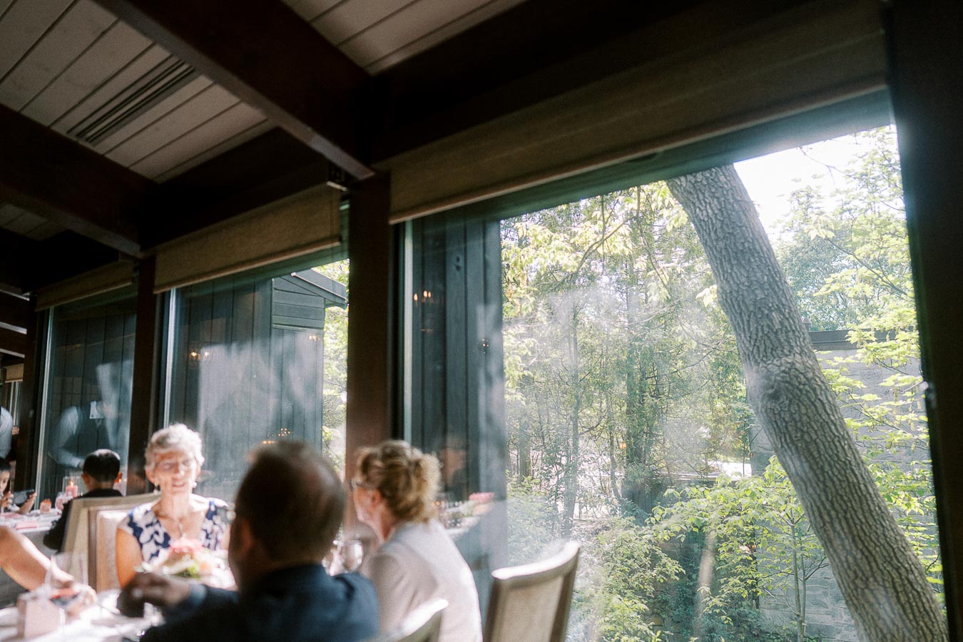 Wedding guests dine beside tall windows overlooking trees during a daytime reception at Ancaster Mill.