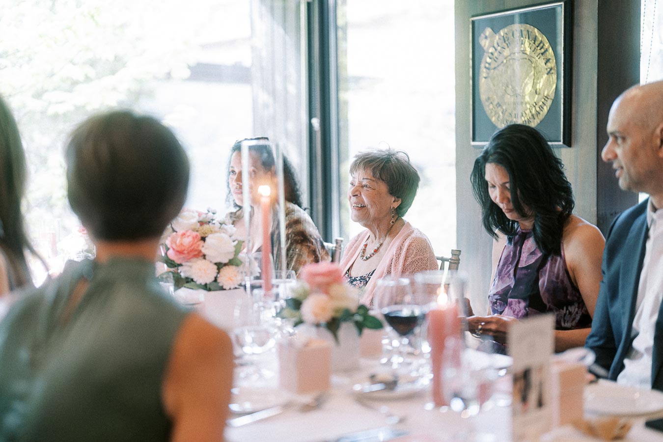 Wedding reception guests seated at a table with pink florals, candles, wine glasses, and soft window light.
