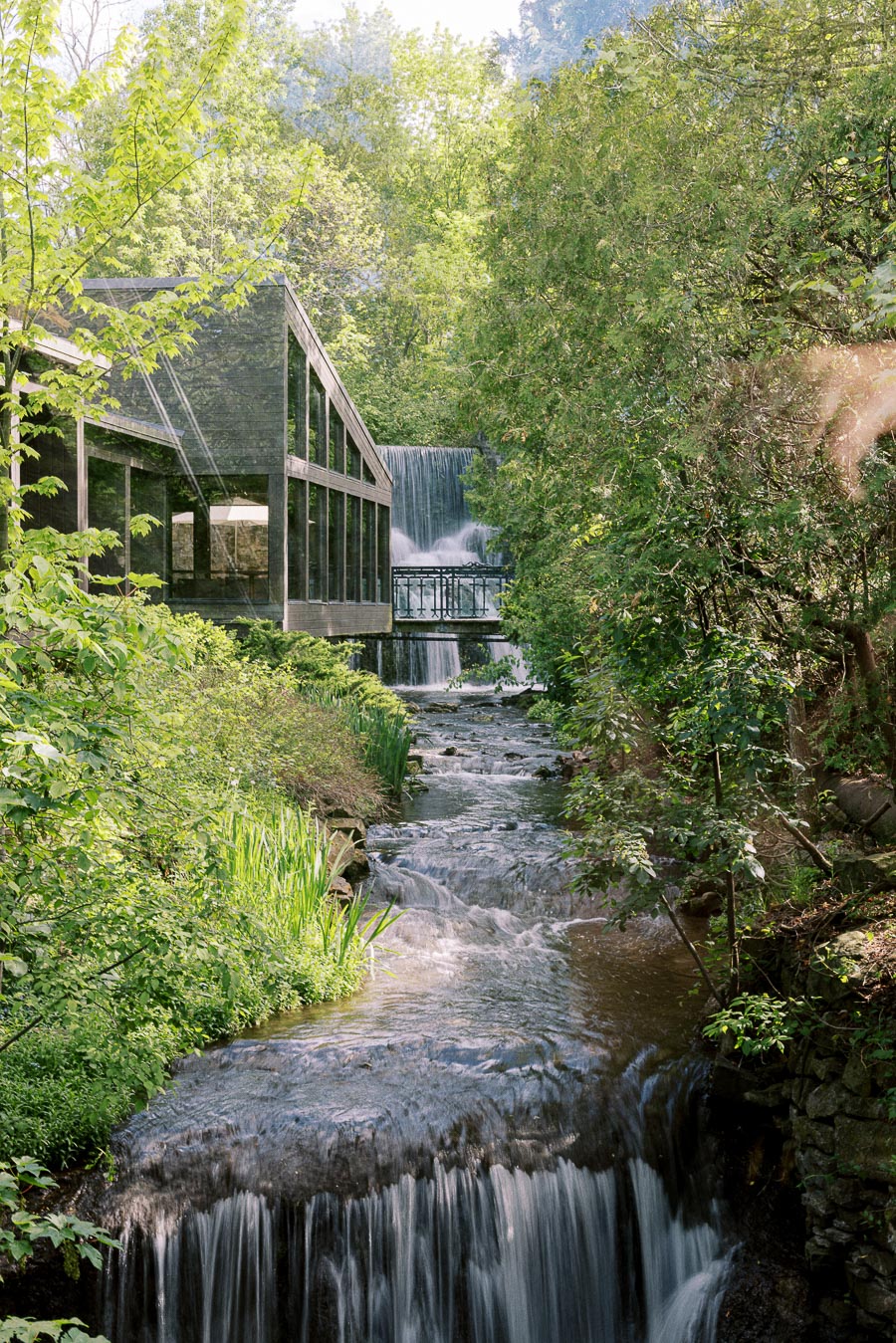 Waterfall and creek flowing beside the glass building and bridge at Ancaster Mill in a wooded setting.