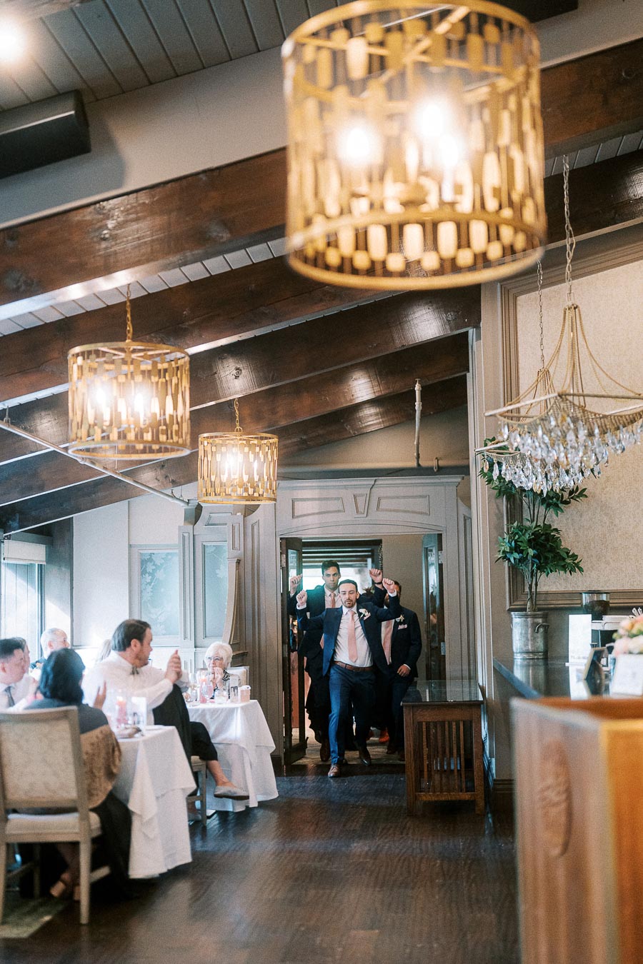 Groom and wedding party enter a chandelier-lit reception room as seated guests watch and clap.