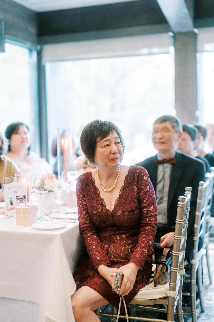 Wedding guests seated at a reception table, with a woman in a burgundy lace dress in the foreground.