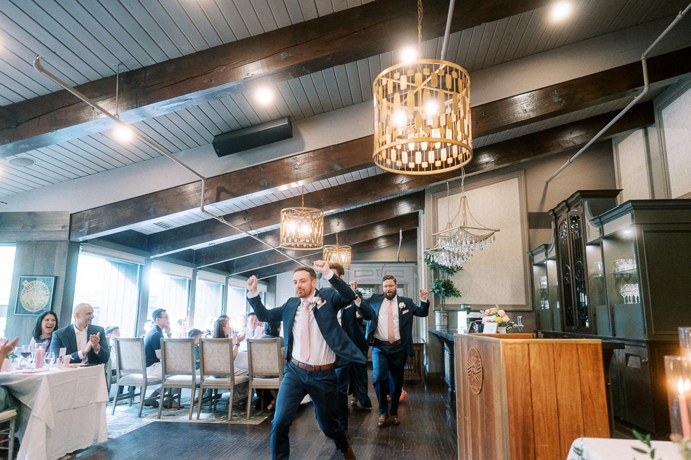 Groomsmen in navy suits dance into a rustic wedding reception room as seated guests clap and cheer.