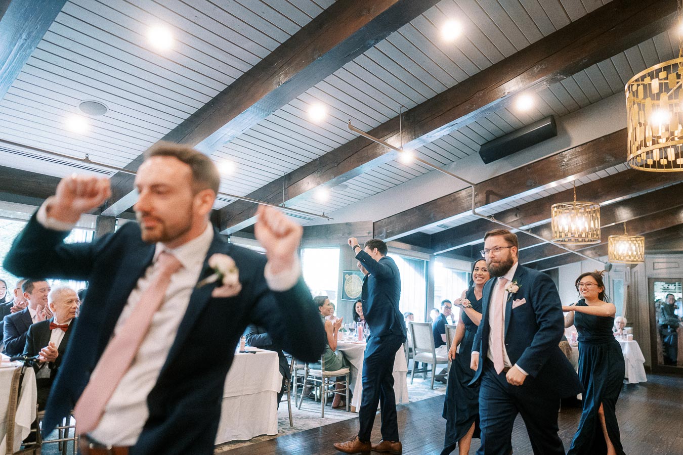 Wedding party members in navy suits and dresses dancing through the reception room beneath wood beams and chandeliers.
