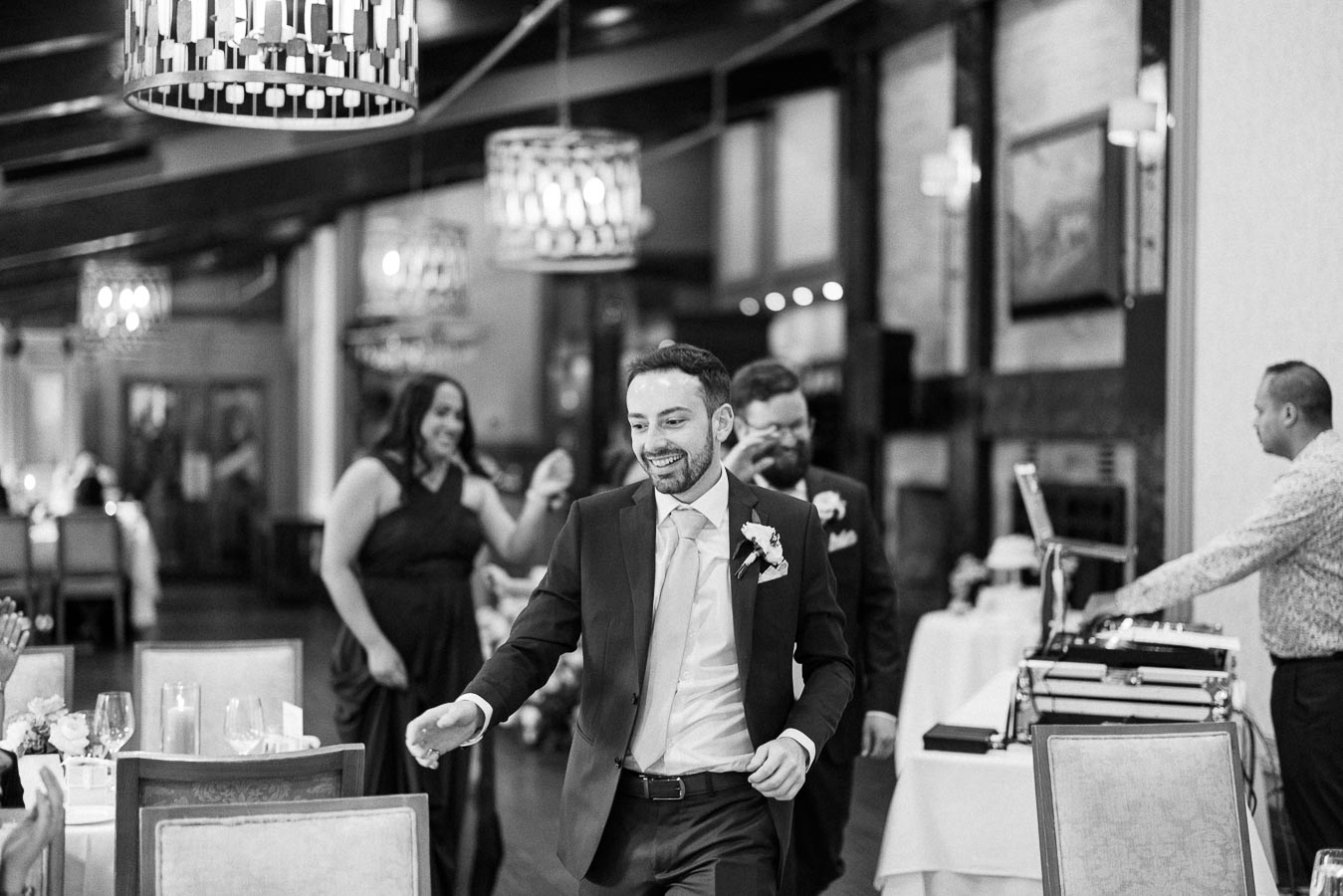 Groom dances between reception tables near the DJ booth while wedding guests smile in the background.