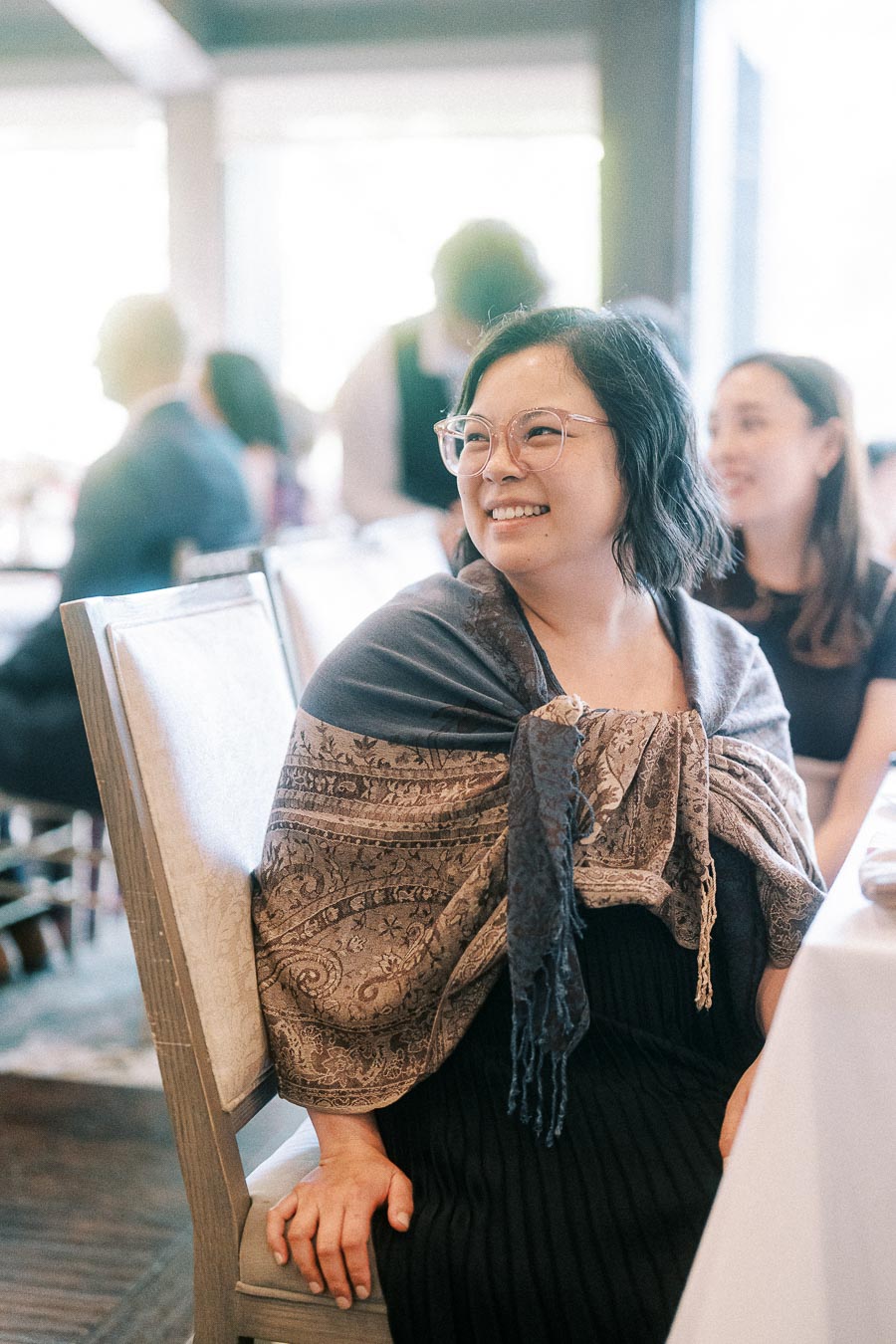 Smiling wedding guest with glasses seated at a reception table, wrapped in a patterned shawl.