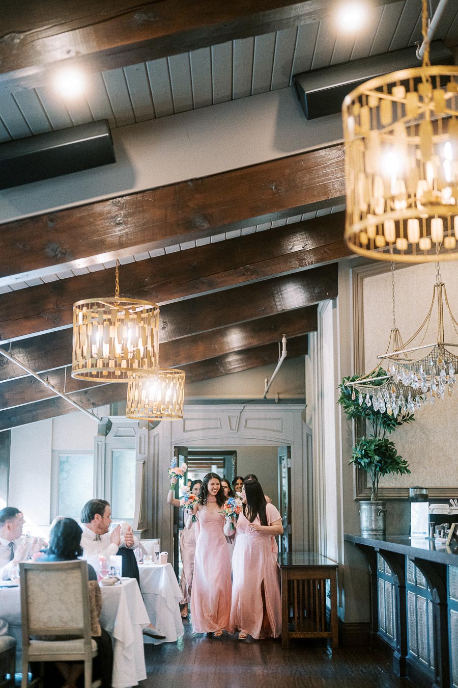 Bridesmaids in blush pink dresses enter a chandelier-lit dining room with guests seated at tables.