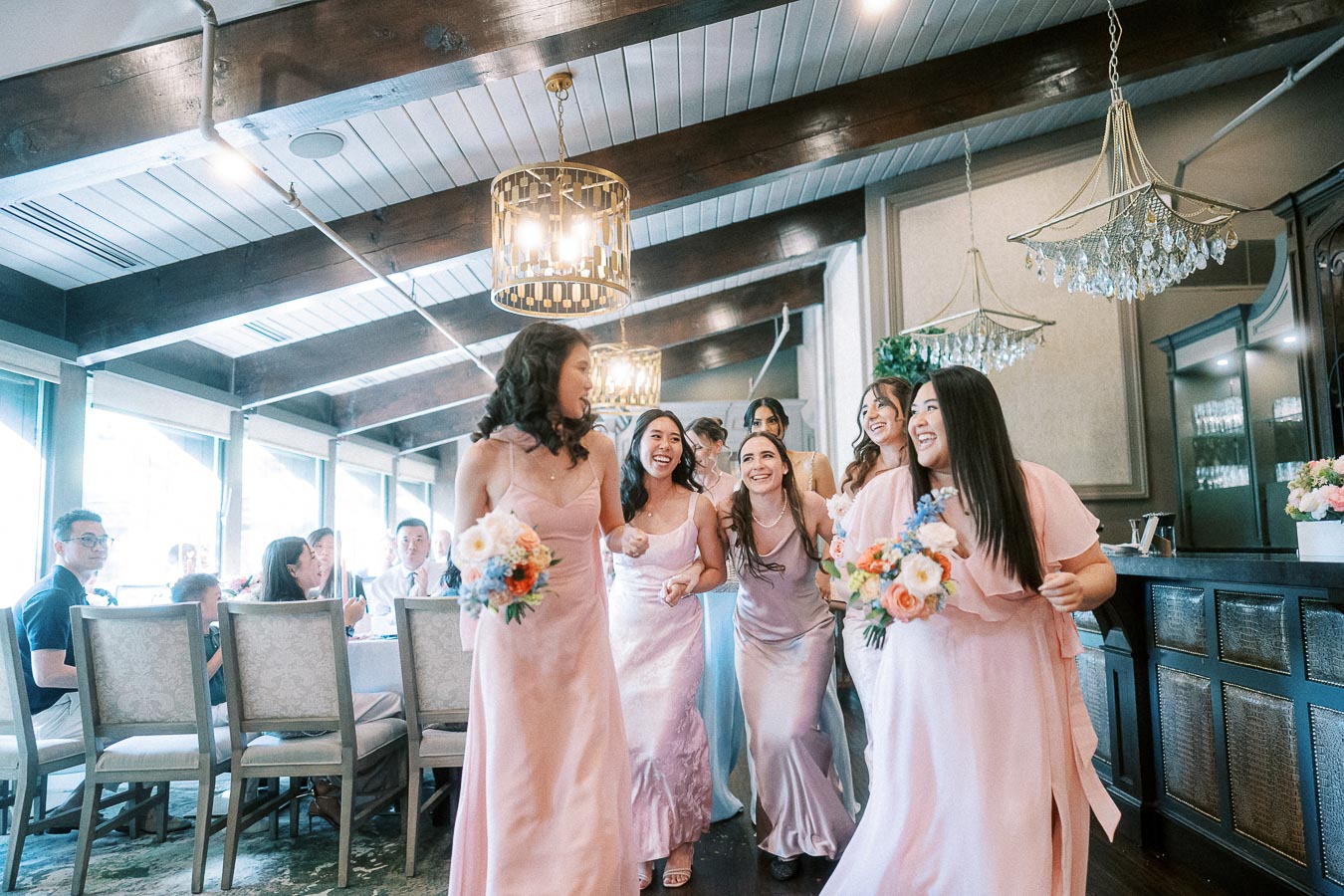 Bridesmaids in blush pink dresses laugh together while holding bouquets in an elegant reception room.