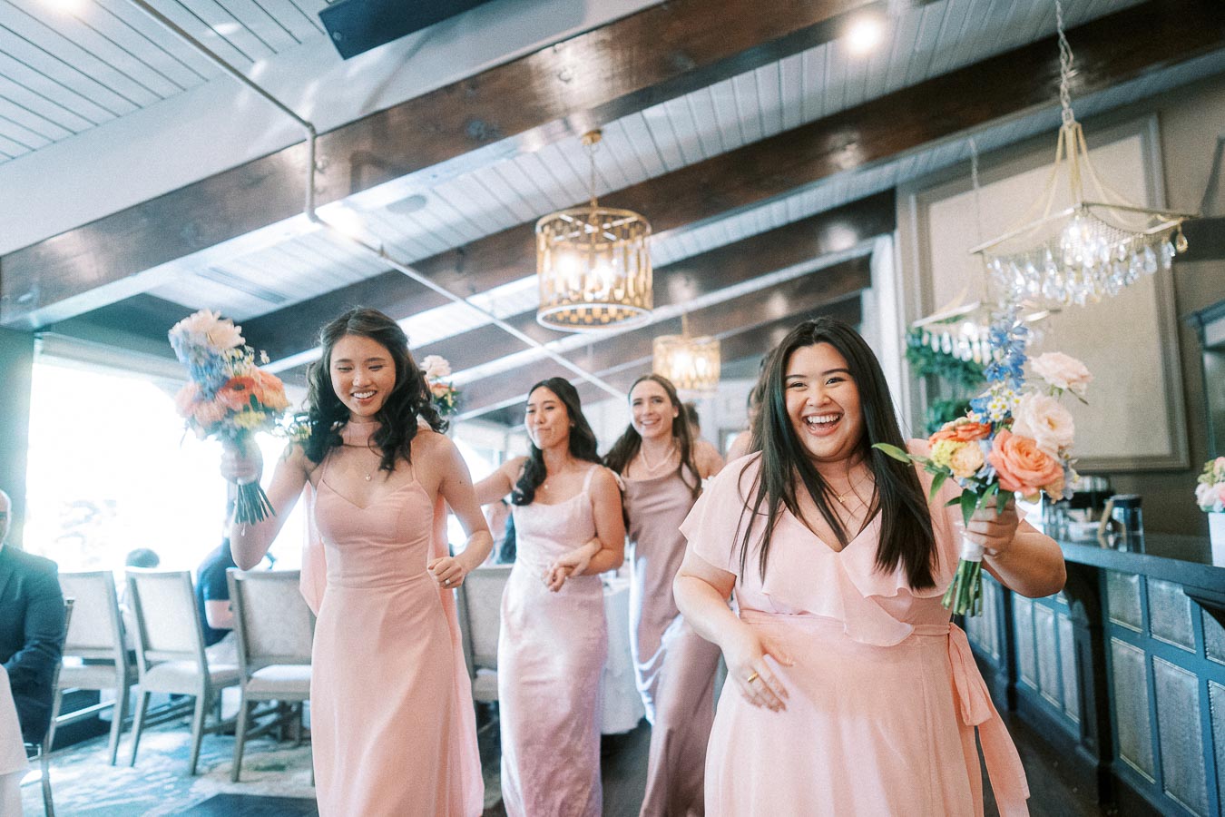 Bridesmaids in blush pink dresses walk indoors carrying pastel bouquets under chandeliers at a wedding reception space.