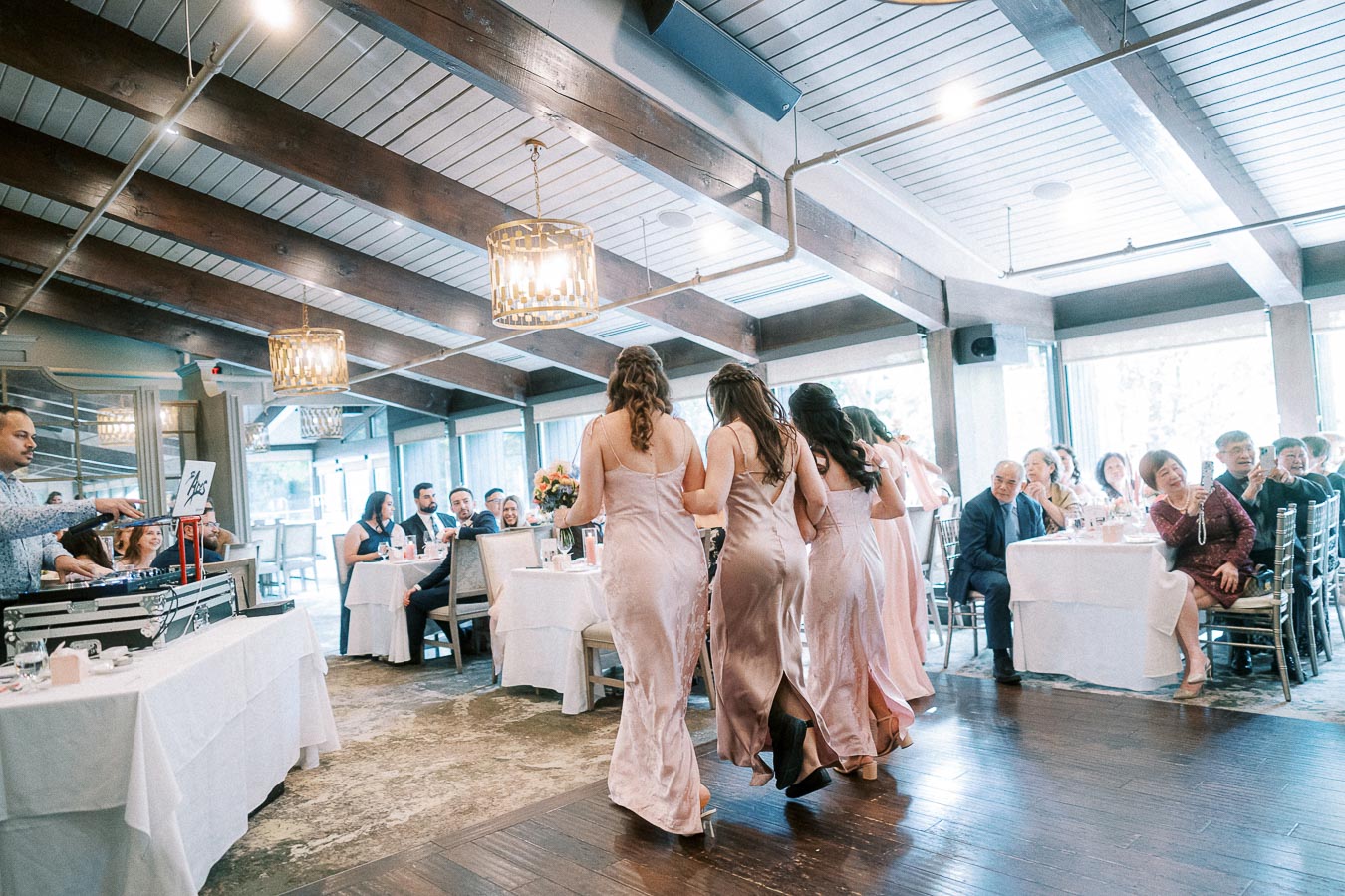 Bridesmaids in pale pink dresses walk together through a bright wedding reception room with seated guests.