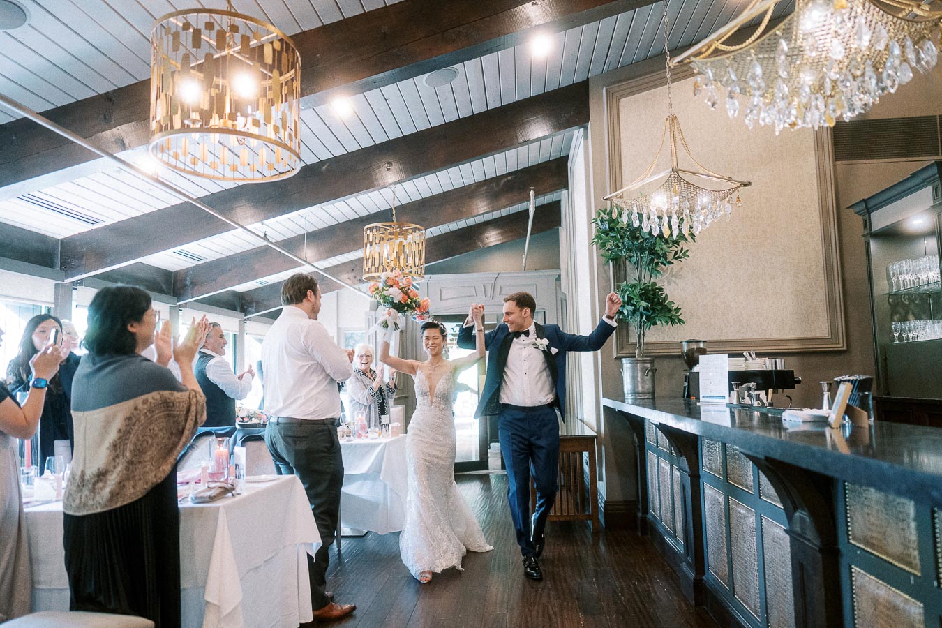 Bride and groom enter their reception holding hands as guests applaud beside a bar and chandeliers.