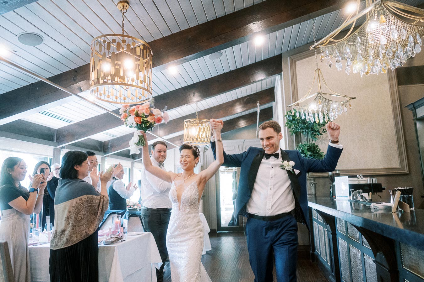 Bride and groom enter a chandelier-lit reception room with raised arms as guests clap beside dining tables.