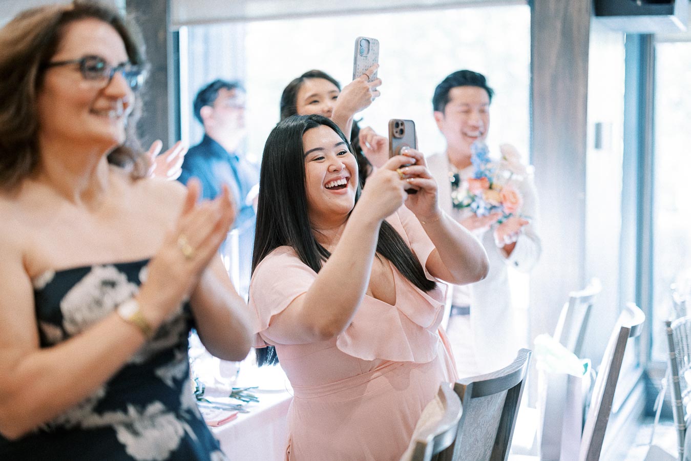 Smiling wedding guests stand indoors clapping and recording on phones during a reception moment.
