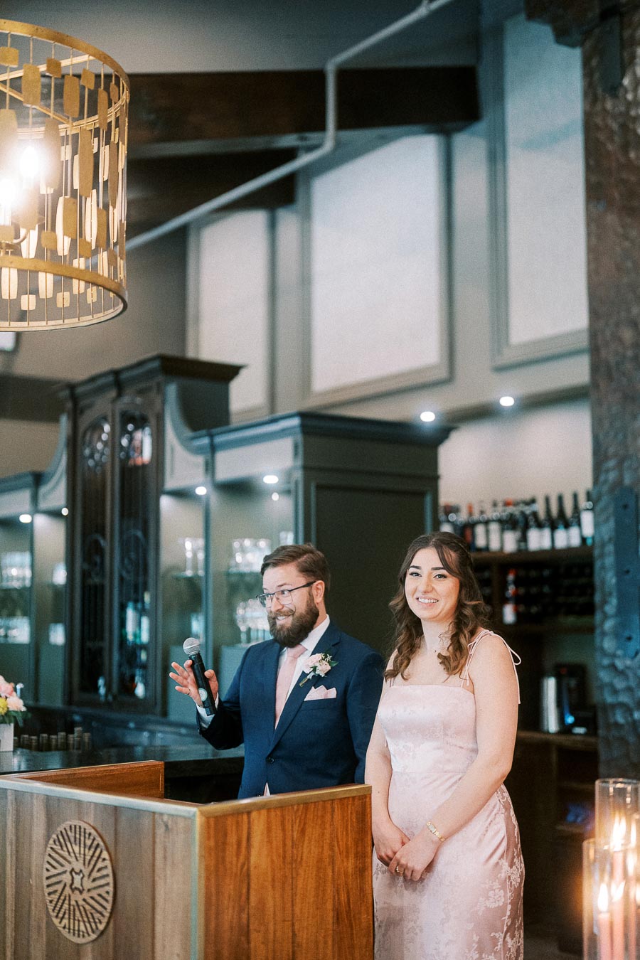 Man in a navy suit holds a microphone beside a woman in a pink dress at a wooden podium in a wedding reception bar.