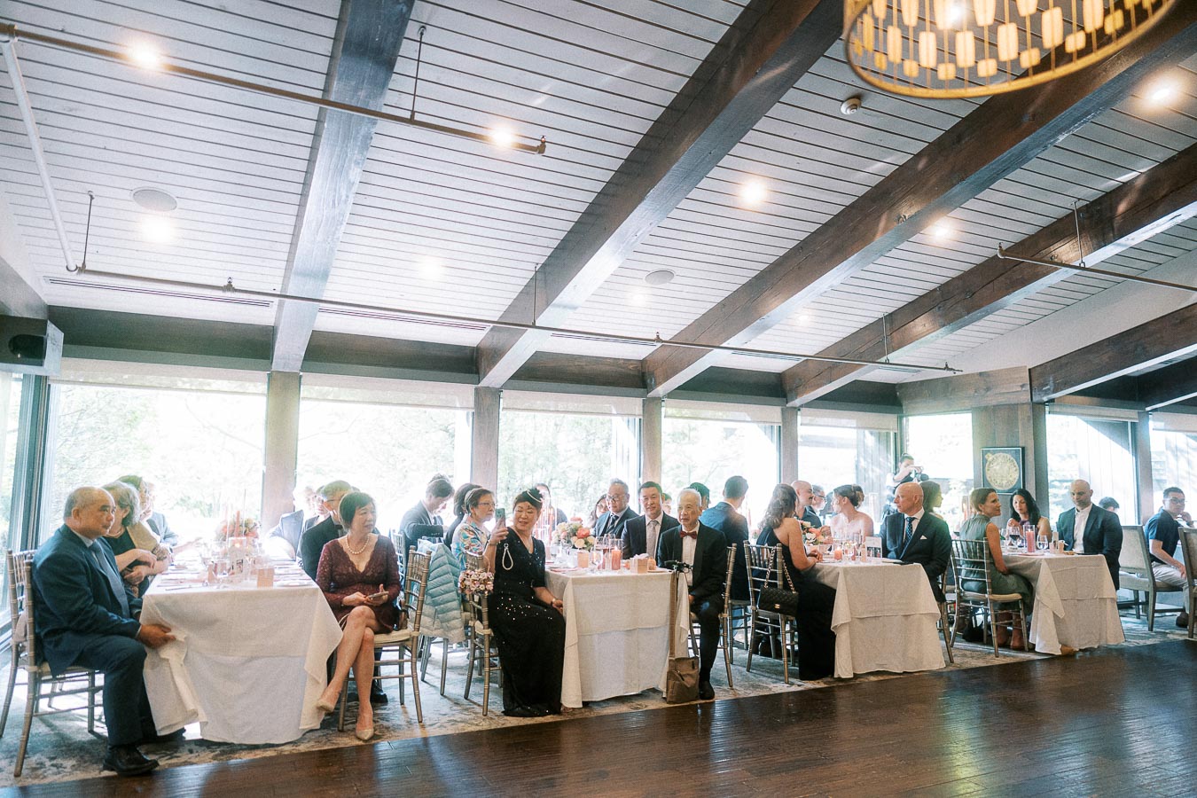 Wedding guests seated at round reception tables in a bright room with wood beams and large windows.