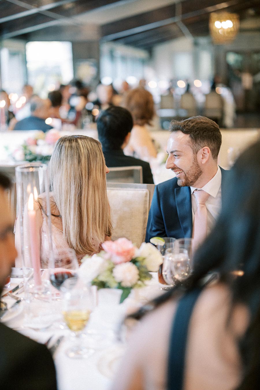Wedding guests talk and laugh at a candlelit reception table with flowers and drinks.