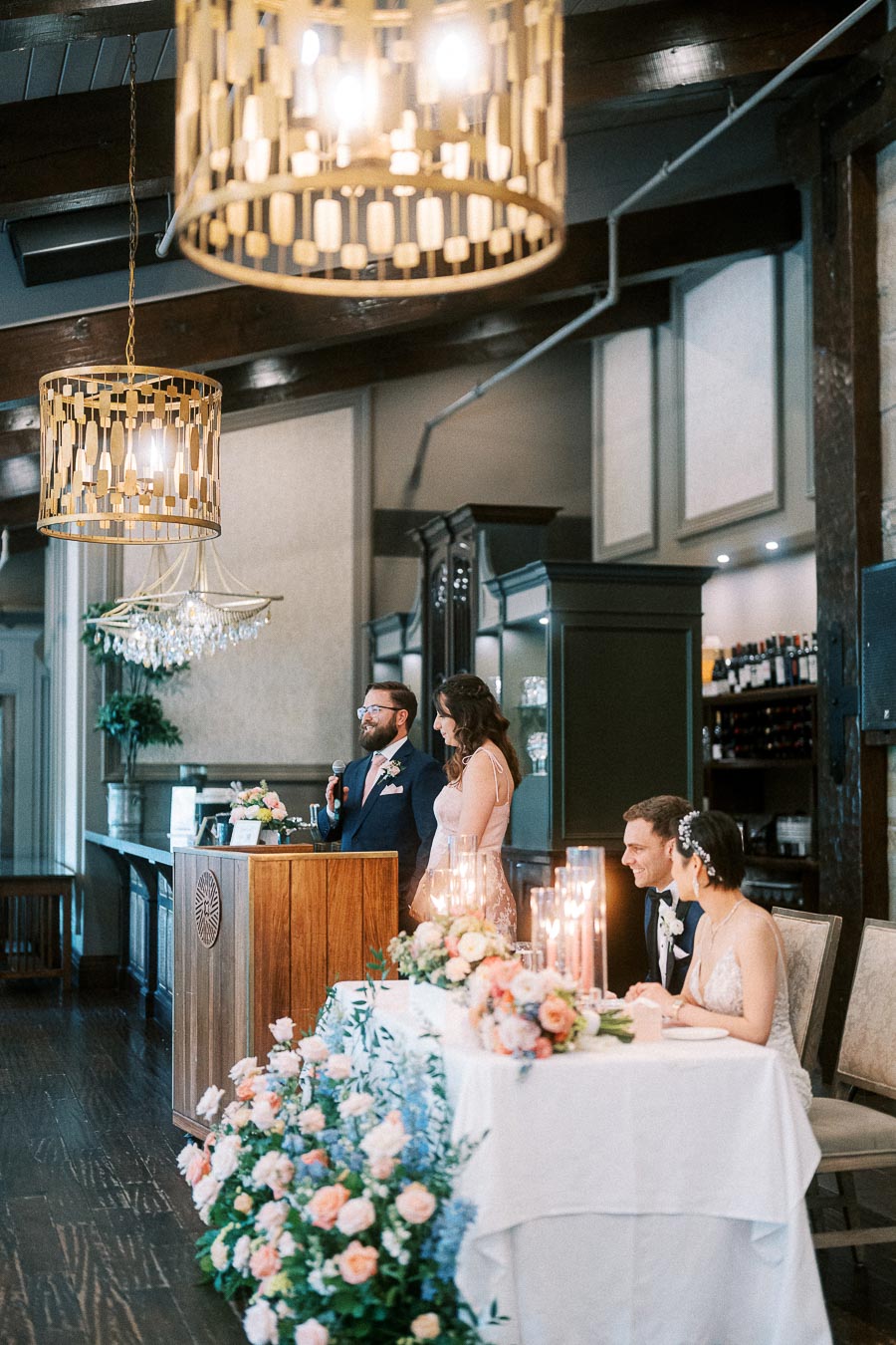 Wedding speaker at a wooden podium while Miffy and Rawle sit at the floral head table beneath chandeliers.