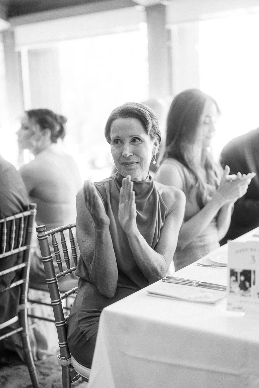 Wedding guest seated at a reception table applauding beside other guests in a bright dining room.