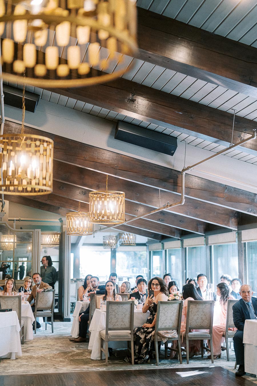 Wedding reception guests seated at tables beneath chandeliers and wood beams at Ancaster Mill.