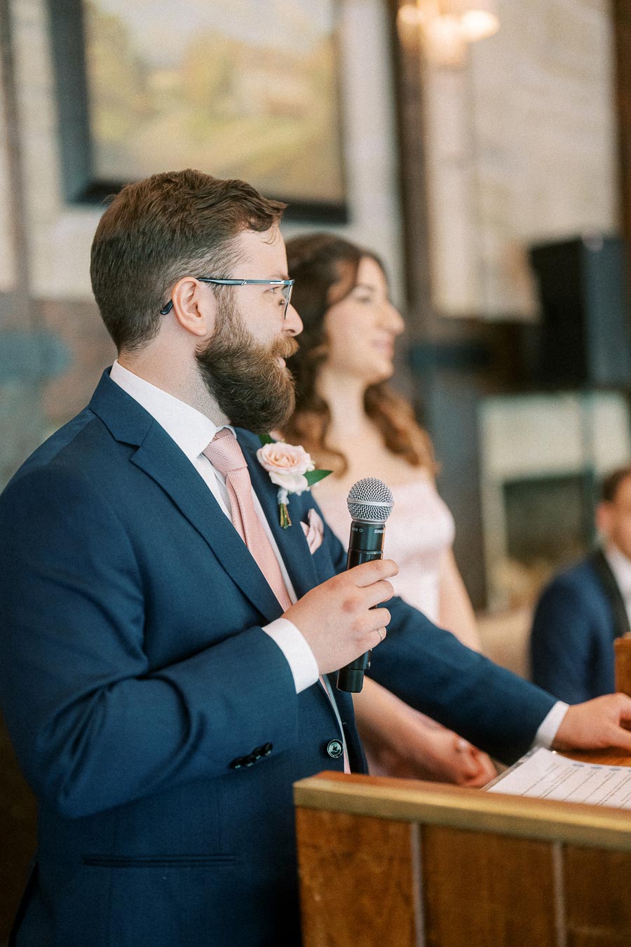 Bearded man in a navy suit holds a microphone at a podium with a woman standing behind him indoors.